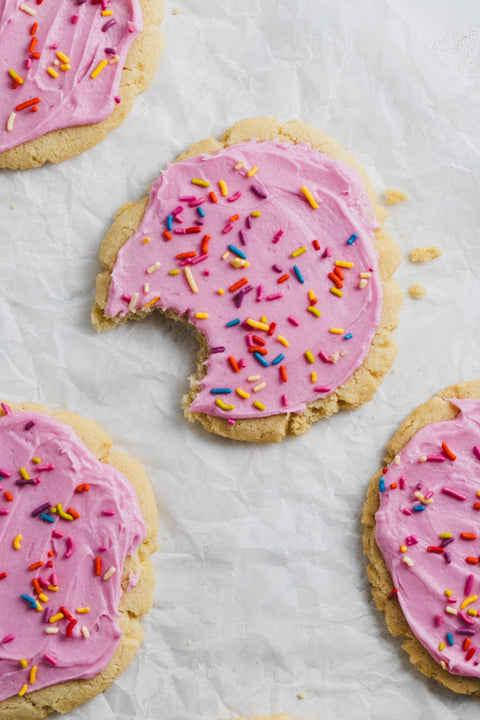 A set of pink frosted sugar cookies with rainbow sprinkles on top, and one half-eaten cookie showing the creamy pink icing and sprinkles.