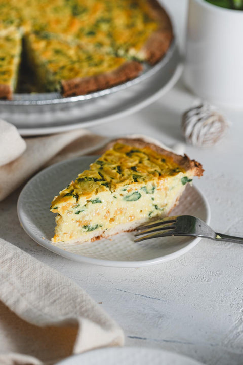 A slice of vegan quiche on a white plate with a fork and the rest of the quiche in the background
