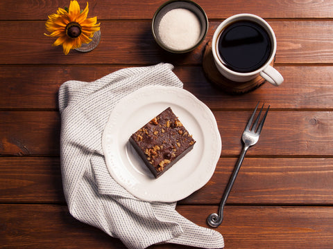 Single walnut brownie on a white plate with a fork, a cup of coffee, a bowl of sugar, and a sunflower on a wooden table.