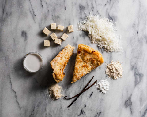 Two slices of coconut custard pie with ingredients including coconut flakes, tofu cubes, vanilla beans, and flour on a marble surface.