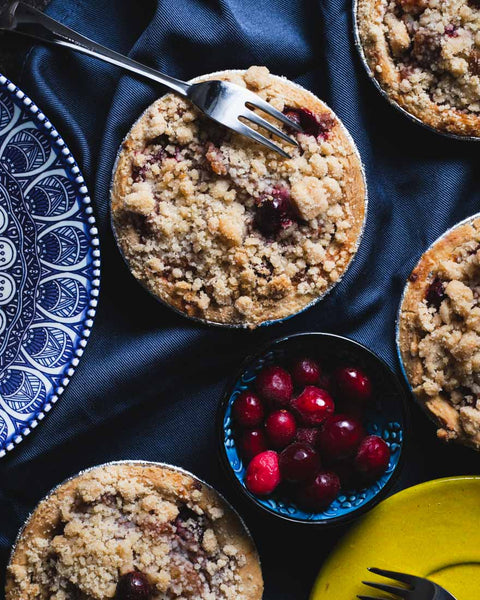 Mini cranberry apple pie with a crumb topping, served with a fork on a dark blue cloth, surrounded by fresh cranberries.