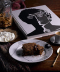 Breakfast protein bar served on a white plate with a cup of espresso and a dollop of whipped cream. The meal is accompanied by a jar of granola and a stylish black-and-white book on a wooden table.
