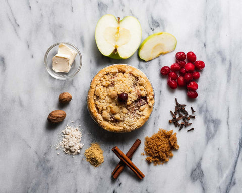 Mini cranberry apple pie with ingredients: apple slices, cranberries, nutmeg, flour, butter, cinnamon sticks, and brown sugar on a marble surface.