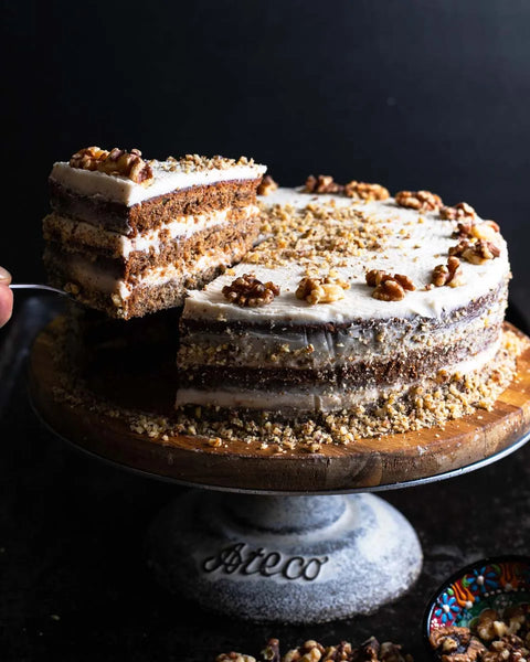 Layered banana walnut cake topped with walnuts and finely chopped nuts, placed on a cake stand with a slice being lifted to reveal the moist interior.
