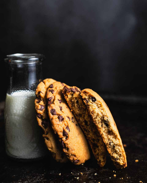 Three chocolate chip cookies leaning against a small glass bottle of milk, with one cookie broken to show the inside, set on a dark surface.