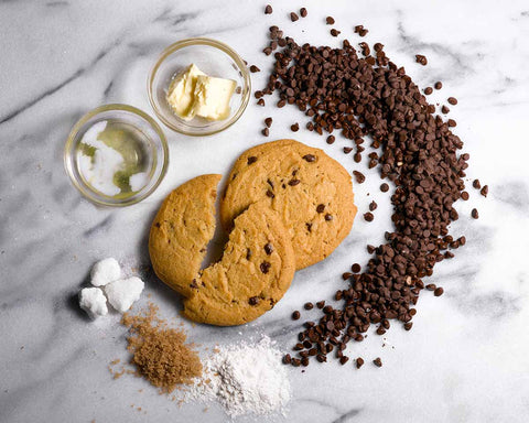 Ingredients for chocolate chip cookies, including butter, oil, flour, brown sugar, chocolate chips, and three baked cookies, arranged on a marble surface.