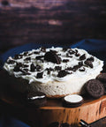 Whole Oreo cheesecake topped with whipped cream and crushed Oreo cookies, placed on a wooden cake stand with whole and half Oreo cookies scattered around.