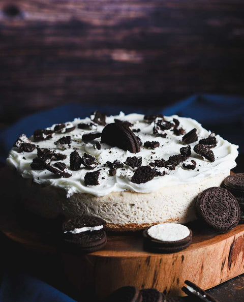 Whole Oreo cheesecake topped with whipped cream and crushed Oreo cookies, placed on a wooden cake stand with whole and half Oreo cookies scattered around.