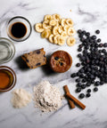 Banana blueberry muffin surrounded by blueberries, bananas, and cinnamon sticks on a white marble background.