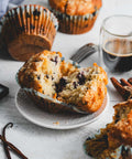Close-up of a blueberry crumb muffin, broken in half to reveal the moist interior with visible blueberries, set on a small place surrounded by a cup of coffee and more muffins in the background.