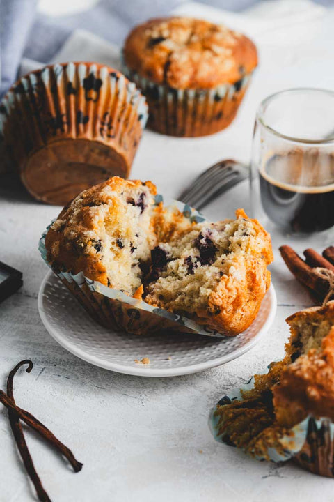 Close-up of a blueberry crumb muffin, broken in half to reveal the moist interior with visible blueberries, set on a small place surrounded by a cup of coffee and more muffins in the background.
