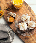 Three carrot walnut muffins with creamy frosting and crushed walnuts on a glass plate, accompanied by a sliced muffin, a glass of orange juice, a yellow flower, and a grey napkin on a wooden board.