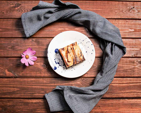 Cheesecake brownie on a white plate with a decorative drizzle, placed on a wooden table with a grey cloth and a pink flower.
