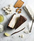 Ingredients for chocolate cheesecake, including tofu, vegan cream cheese, lemon, vanilla bean, flour, sugar, chocolate, and vegetable oil, arranged around a slice of cheesecake on a marble surface.