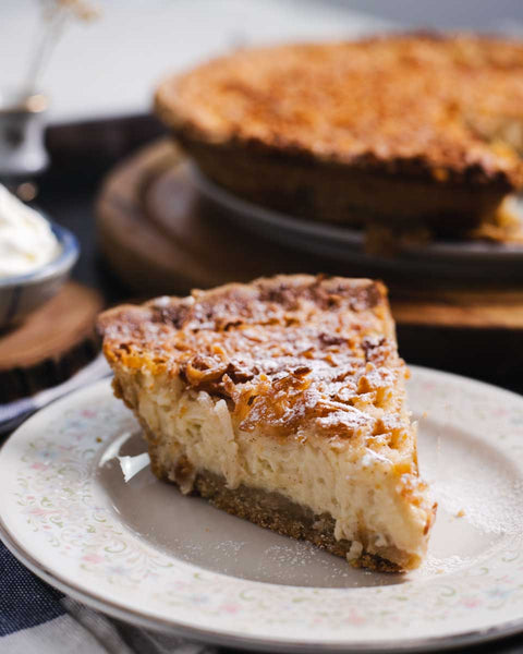 Slice of coconut custard pie on a decorative plate, with the rest of the pie in the background on a wooden stand.