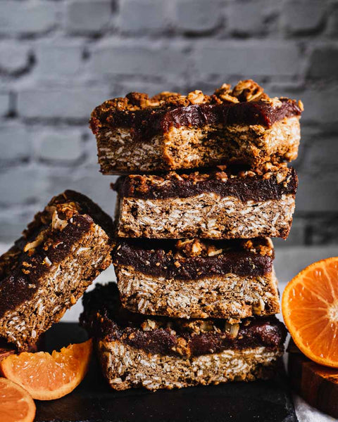 Stack of orange date bars with layers of oats and date filling, with fresh orange slices on a dark surface and a brick wall background.