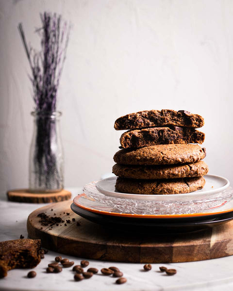 Stack of espresso fudge cookies on a plate, with a broken cookie on top, on a wooden surface with coffee beans scattered around.