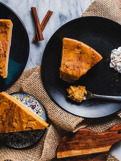 Top-down view of two slices of pumpkin cheesecake on black plates, with cinnamon sticks and a dollop of whipped cream on a marble surface and burlap cloth.