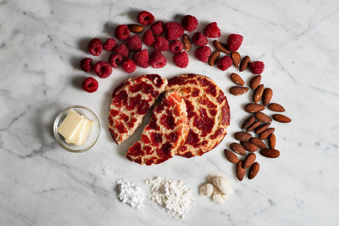 Ingredients for raspberry crinkle cookies including fresh raspberries, almonds, butter, and flour, arranged around sliced cookies on a marble surface.