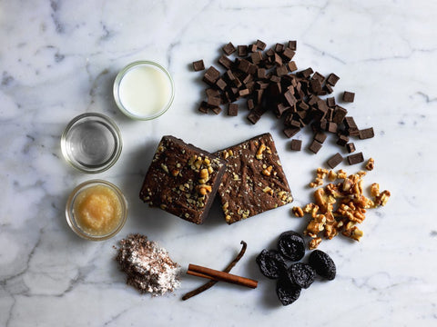 Ingredients for walnut brownie on a marble surface, including chocolate chunks, walnuts, prunes, milk, oil, applesauce, flour, cinnamon sticks, and vanilla beans, with two walnut brownies in the center.