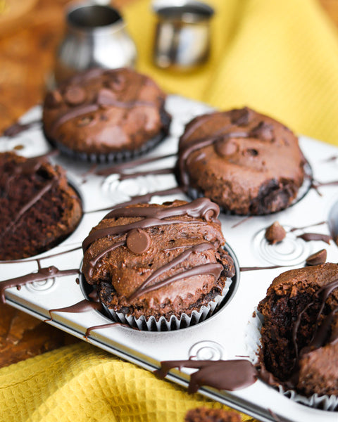 Cocoa bean muffins drizzled with chocolate in a muffin tin, displayed on a yellow cloth.