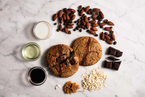 Two espresso fudge cookies with ingredients: almonds, coffee beans, chocolate chunks, oats, brown sugar, and oil on a marble surface.