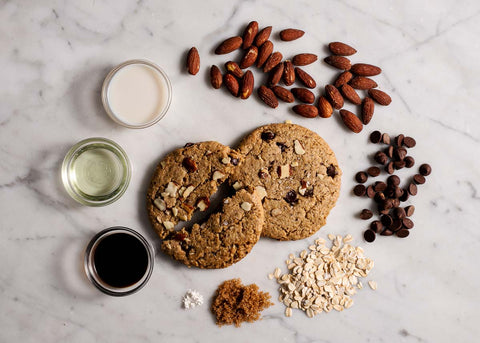 Top view of two salted almond chocolate chip cookies surrounded by ingredients: almonds, chocolate chips, oat flour, sugar, oil, vanilla, and milk on a marble surface.