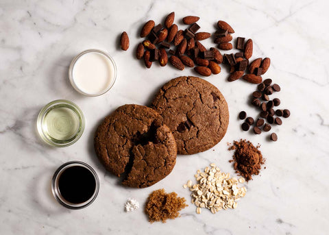 Two milk chocolate cookies on a marble surface, surrounded by ingredients including almonds, chocolate chips, oats, cocoa powder, brown sugar, milk, vanilla extract, and oil.