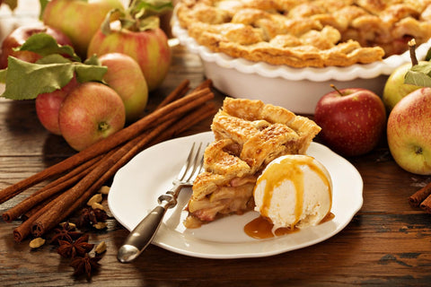 A slice of apple pie on a white plate with vanilla ice cream surrounded by cinnamons sticks and apples