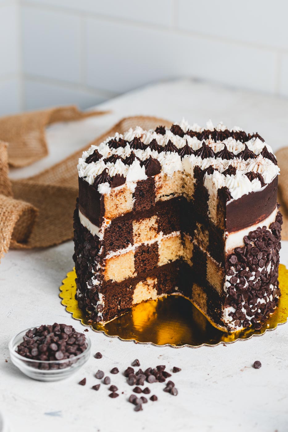 A cake with elaborate chocolate and vanilla striped decorative icing rolled in chocolate chips and lightly dusted with cocoa powder. It is cut open showing a chocolate and vanilla checkerboard patterned cake inside and there is a bowl of chocolate chips in front of it