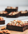 A sugar free keto brownie with a crushed almond topping on a wooden tray with almonds scattered around and more brownies in the background