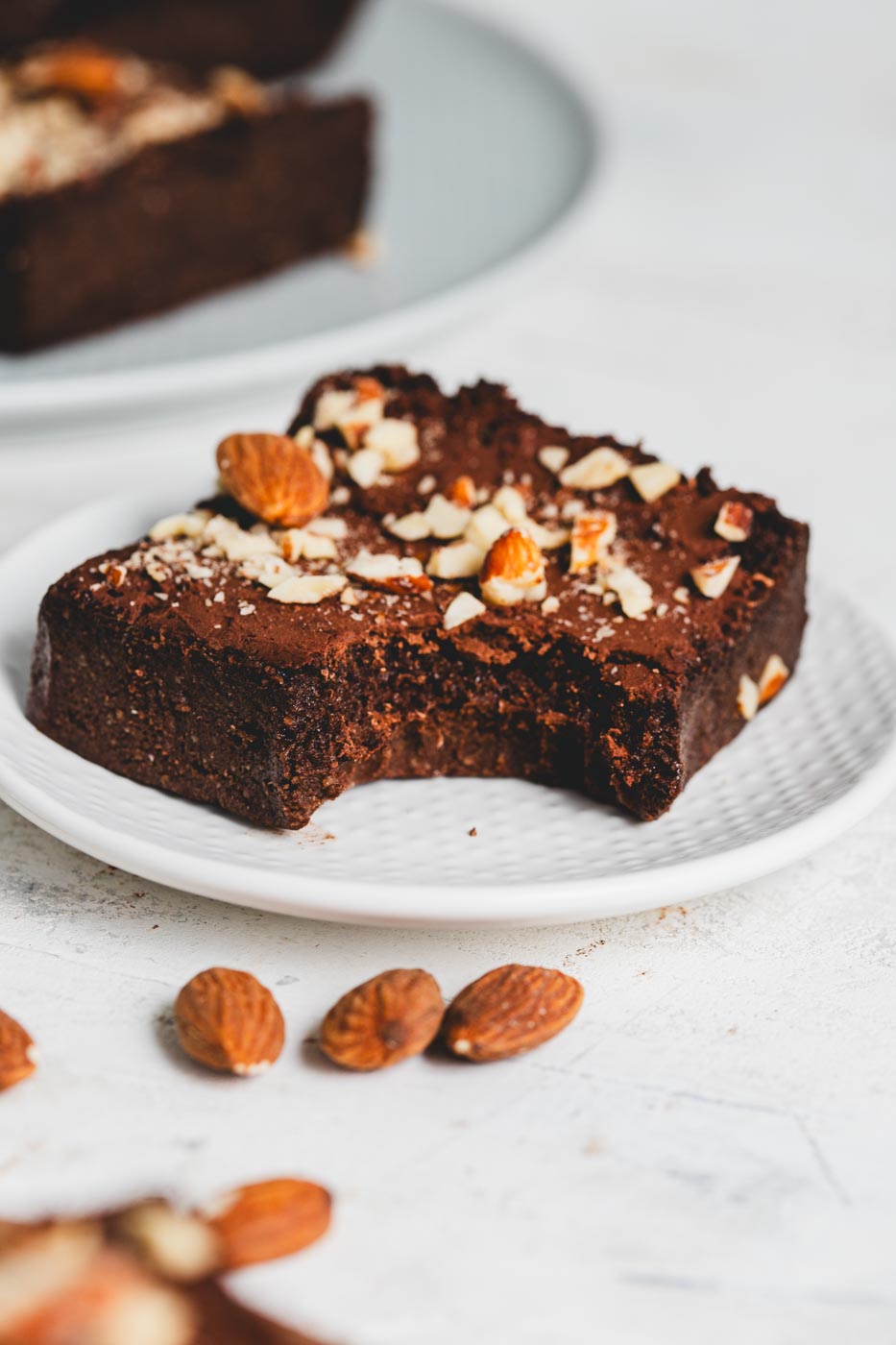 A sugar free keto brownie with a crushed alomond topping with a bite taken out of it to show fudgey texture on a white plate  with almonds scattered around