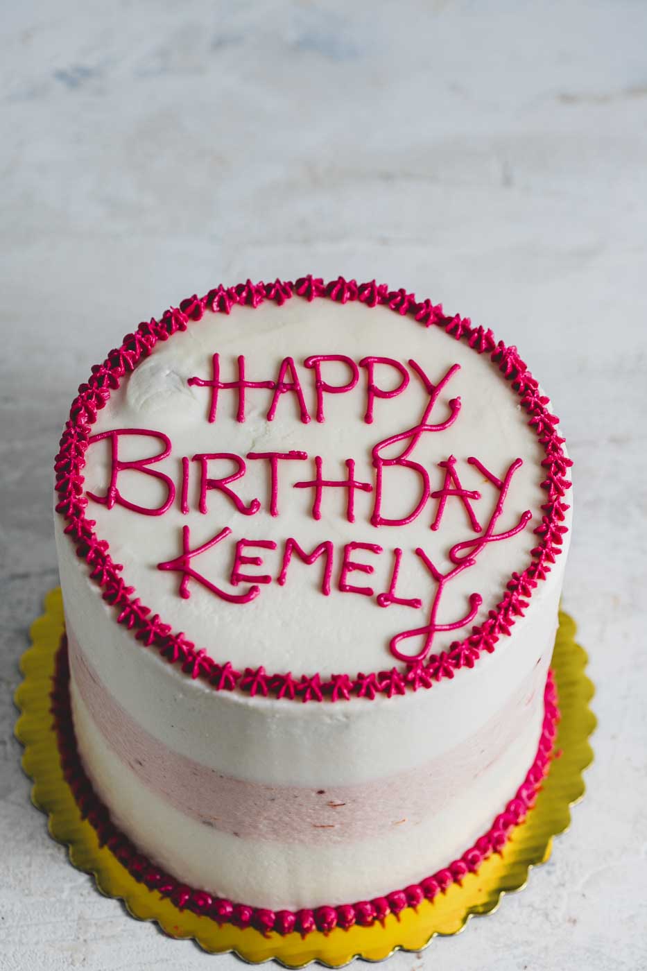 A cake with striped vanilla and strawberry frosting with bright pink frosting decorations and "happy birthday kemely" written on top