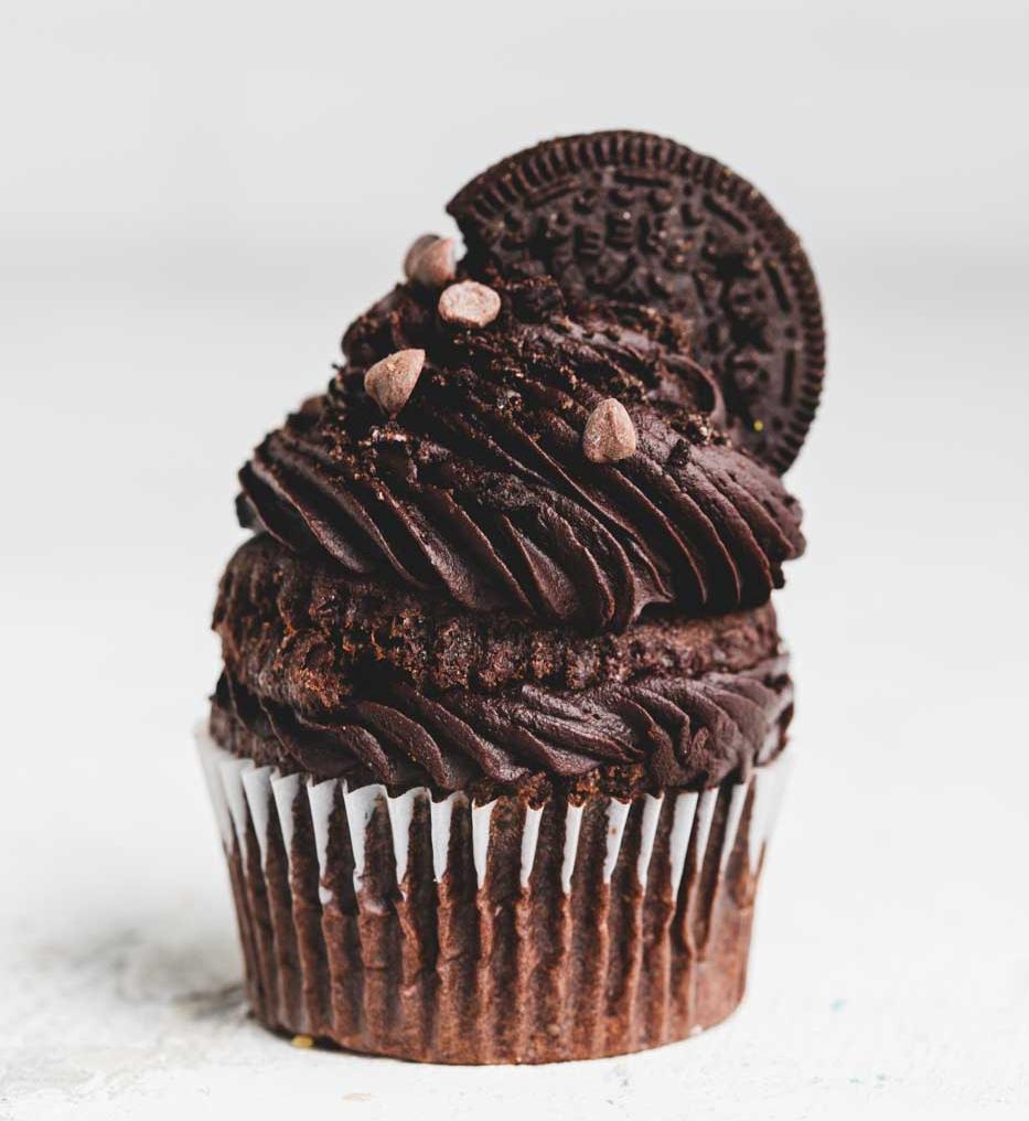 A close up of a chocolate cupcake with chocolate icing and chocolate chips with an oreo cookie on top 