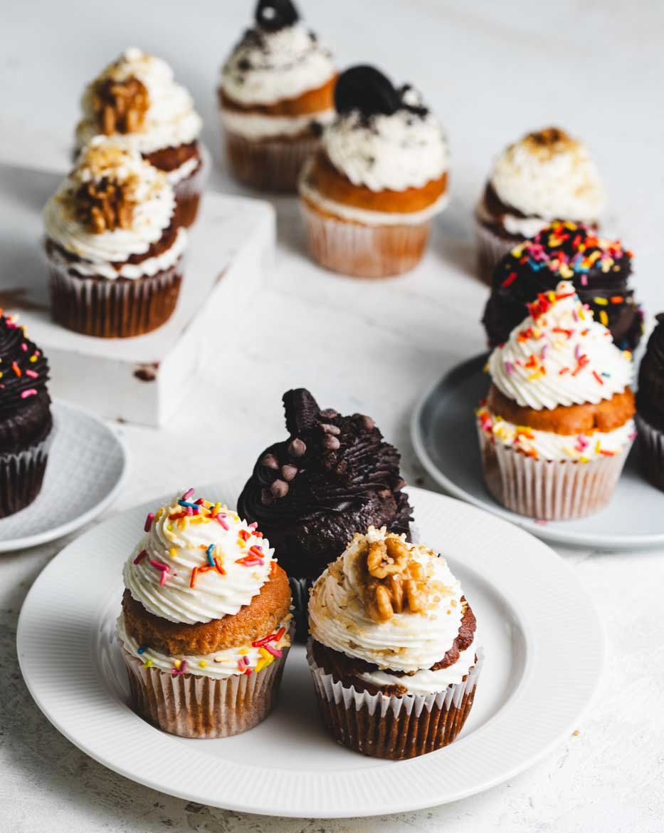 White plates of chocolate, vanilla, and banana walnut cupcakes with chocolate and vanilla frosting topped with rainbow sprinkles, walnuts, chocolate chips, and oreo cookies.
