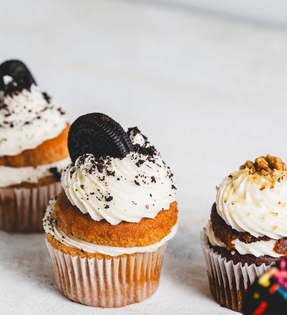 A close up of a vanilla cupcake with vanilla icing and an oreo cookie garnish with more vanilla and banana walnut cupcakes around it
