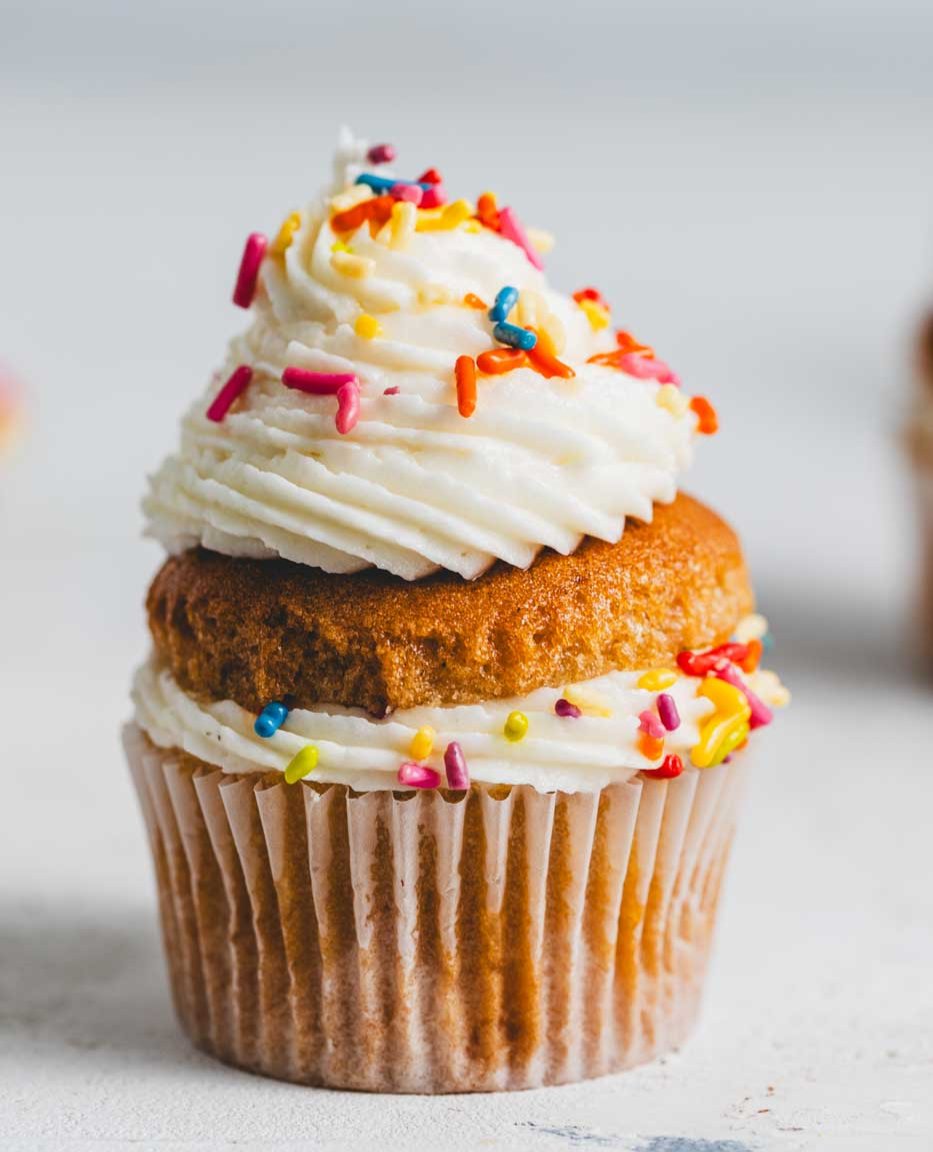 A close up of a vanilla cupcake with vanilla icing and rainbow sprinkles