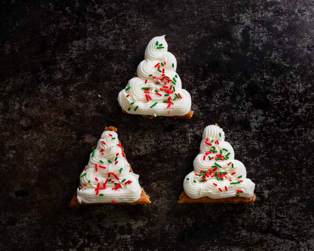 Three Christmas tree-shaped cookies with white frosting and red and green sprinkles, arranged on a dark rustic surface.