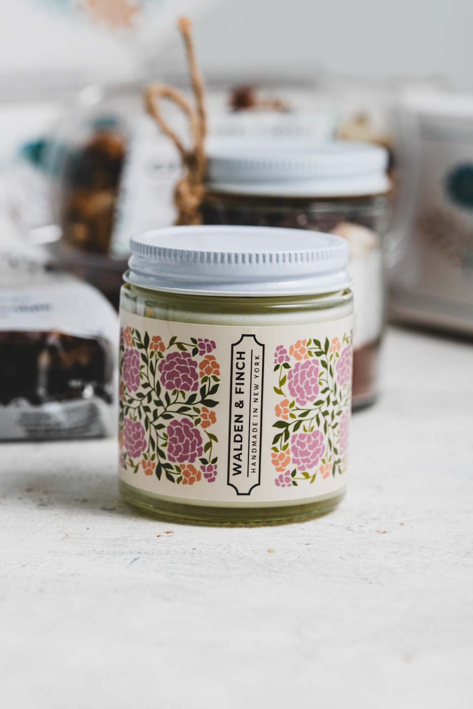 close up of a walden & finch handmade jar candle with various treats and an open gift box in the background