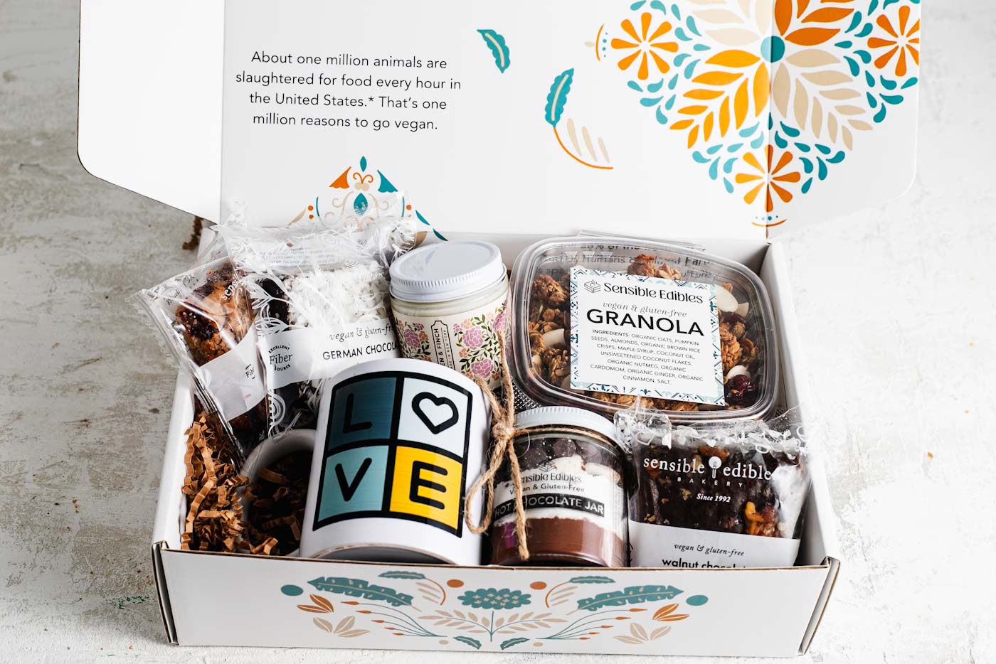 Gift box opnened to show contents: a jar of hot chocolate mix, walnut chocolate brownie, sensible edibles mug, candle, granola, and german chocolate brownie. The box is white with a floral background