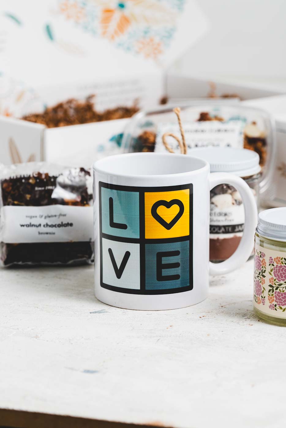 Close up of a sensible edibles mug that says love in blue and yellow squares with various treats and an open gift box in the background