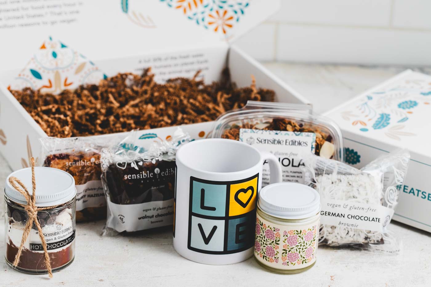 Gift box with contents taken out in front of it: Jar of hot chocolate mix, walnut chocolate brownie, sensible edibles mug, candle, granola, and german chocolate brownie.