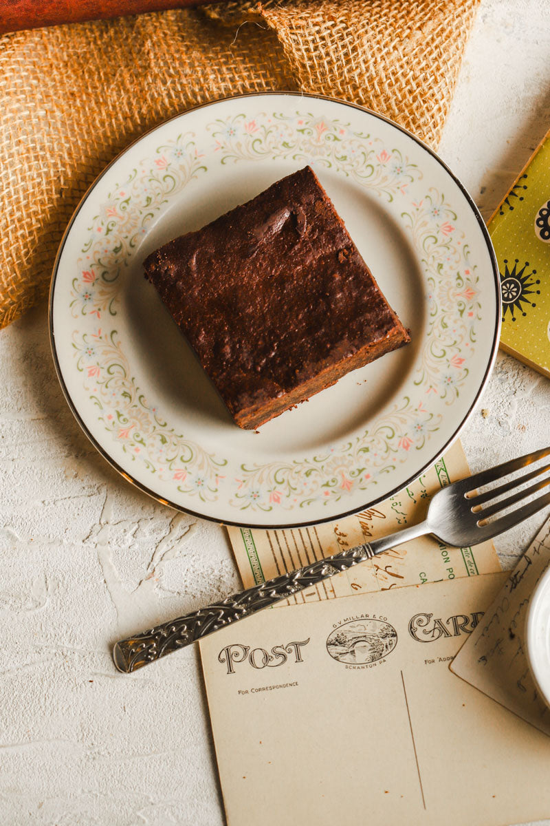 Chocolate chunk brownie on a decorative plate with a fork and postcard in the background
