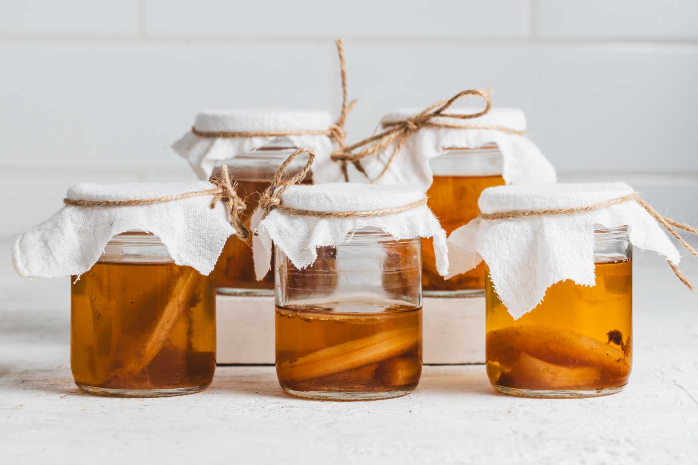 Five small jars of kombucha with scobies floating inside lined up on a white background