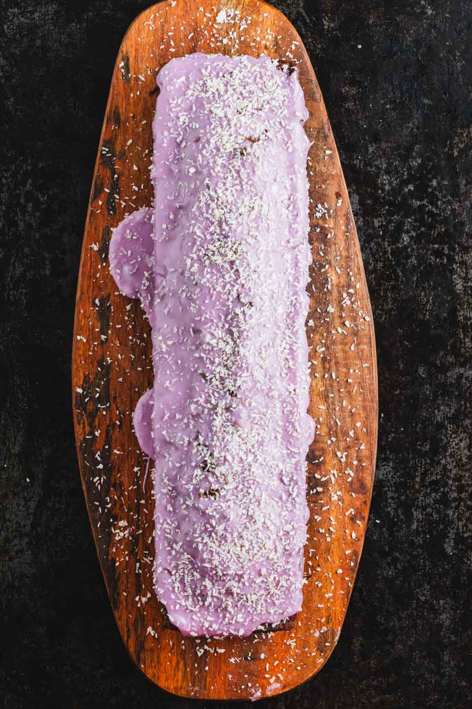 A whole blueberry coconut loaf with a vibrant purple blueberry icing and coconut flake garnish seen from above on a wooden tray