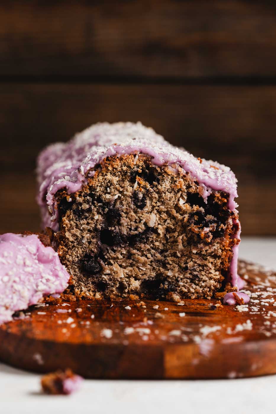 A coconut loaf with blueberries throughout and a vibrant purple blueberry icing garnished with shredded coconut. There is a slice cut off to show the interior of the loaft