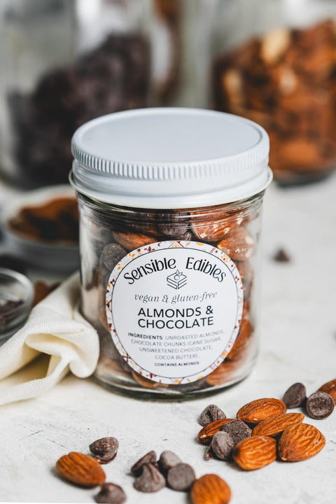 A jar of Sensible Edibles Almonds & Chocolate. The jar contains whole almonds and chocolate chunks, and is surrounded by scattered almonds and chocolate pieces.