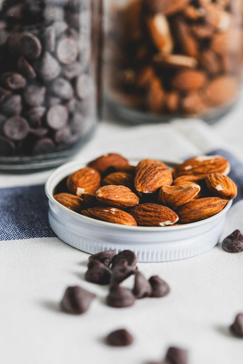 A white jar lid used as a dish holding whole almonds, with scattered chocolate chips around it. Two jars in the background, one filled with chocolate chips and the other with almonds.