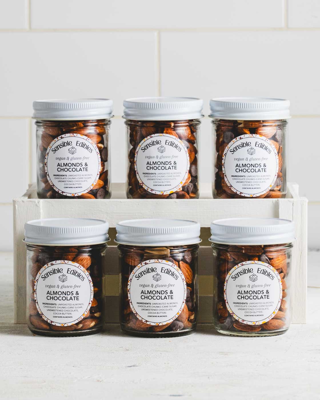 Six jars of Sensible Edibles Almonds & Chocolate with white lids arranged in two rows on a white wooden crate.