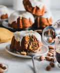 Mini lemon almond bundt cake with white icing and almond slices on a plate, surrounded by whole almonds and a glass of espresso, with more cakes in the background.